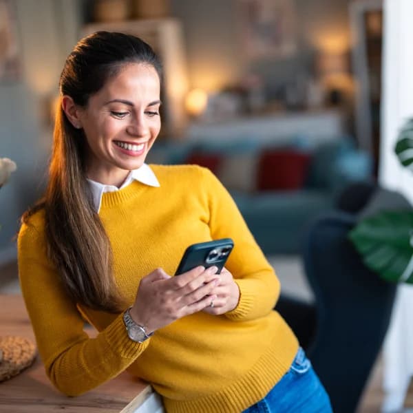 Une femme souriante en pull jaune utilise son téléphone, évoquant un virement Interac.