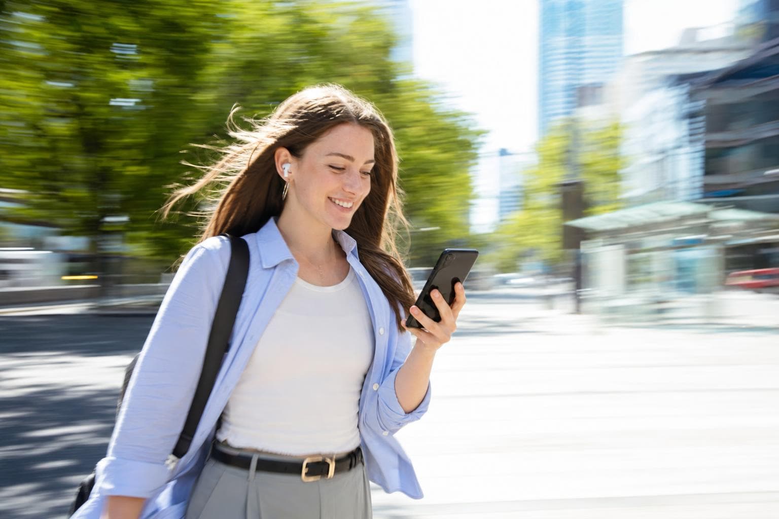 A young woman walks briskly along a bright city street, smiling as she glances at her phone.