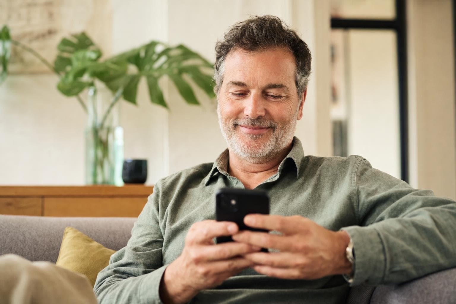 Un homme d'âge moyen, rassuré, assis sur un canapé chez lui, sourit doucement en regardant son téléphone.